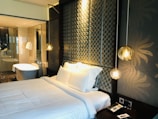 A modern bedroom features a neatly made bed with white linens and several pillows. The headboard has a patterned design with circular motifs, and hanging pendant lights add a warm, ambient glow. The room has an elegant and luxurious feel, complemented by the presence of a bathroom in the background with a standalone bathtub, glass partitions, and white towels hanging nearby. A bedside table has a phone and a small notepad.
