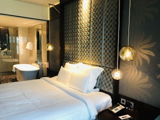 A modern bedroom features a neatly made bed with white linens and several pillows. The headboard has a patterned design with circular motifs, and hanging pendant lights add a warm, ambient glow. The room has an elegant and luxurious feel, complemented by the presence of a bathroom in the background with a standalone bathtub, glass partitions, and white towels hanging nearby. A bedside table has a phone and a small notepad.