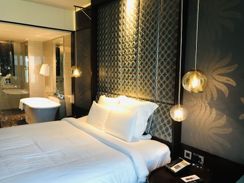 A modern bedroom features a neatly made bed with white linens and several pillows. The headboard has a patterned design with circular motifs, and hanging pendant lights add a warm, ambient glow. The room has an elegant and luxurious feel, complemented by the presence of a bathroom in the background with a standalone bathtub, glass partitions, and white towels hanging nearby. A bedside table has a phone and a small notepad.
