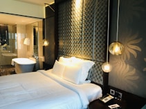 A modern bedroom features a neatly made bed with white linens and several pillows. The headboard has a patterned design with circular motifs, and hanging pendant lights add a warm, ambient glow. The room has an elegant and luxurious feel, complemented by the presence of a bathroom in the background with a standalone bathtub, glass partitions, and white towels hanging nearby. A bedside table has a phone and a small notepad.