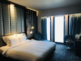 A modern hotel room with a large, comfortable bed featuring white linens and multiple pillows. The headboard has a geometric pattern and integrated ambient lighting. A blue armchair is positioned next to a circular-patterned carpet. Two pendant lights hang beside the bed, and thick blue curtains cover large windows allowing in soft light.