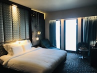 A modern hotel room with a large, comfortable bed featuring white linens and multiple pillows. The headboard has a geometric pattern and integrated ambient lighting. A blue armchair is positioned next to a circular-patterned carpet. Two pendant lights hang beside the bed, and thick blue curtains cover large windows allowing in soft light.