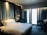 A modern hotel room with a large, comfortable bed featuring white linens and multiple pillows. The headboard has a geometric pattern and integrated ambient lighting. A blue armchair is positioned next to a circular-patterned carpet. Two pendant lights hang beside the bed, and thick blue curtains cover large windows allowing in soft light.