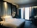 A modern hotel room with a large, comfortable bed featuring white linens and multiple pillows. The headboard has a geometric pattern and integrated ambient lighting. A blue armchair is positioned next to a circular-patterned carpet. Two pendant lights hang beside the bed, and thick blue curtains cover large windows allowing in soft light.