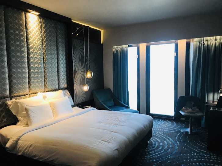 A modern hotel room with a large, comfortable bed featuring white linens and multiple pillows. The headboard has a geometric pattern and integrated ambient lighting. A blue armchair is positioned next to a circular-patterned carpet. Two pendant lights hang beside the bed, and thick blue curtains cover large windows allowing in soft light.