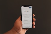 A hand is holding a smartphone displaying the Google search page, which shows trending search topics. The phone screen is bright against a dark background.