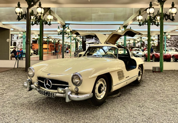 A vintage white Mercedes-Benz sports car is displayed in an elegant showroom. The car features iconic gull-wing doors that are open, showcasing its luxurious design. The background has ornate green lamp posts and mirrored walls reflecting other classic cars and visitors, creating an atmosphere of sophistication and nostalgia.
