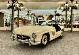 A vintage white Mercedes-Benz sports car is displayed in an elegant showroom. The car features iconic gull-wing doors that are open, showcasing its luxurious design. The background has ornate green lamp posts and mirrored walls reflecting other classic cars and visitors, creating an atmosphere of sophistication and nostalgia.