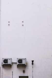 A large, plain white wall with visible cracks and two air conditioning units mounted side by side. There is a lamp fixture and a few small vents on the wall.