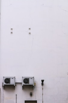 A large, plain white wall with visible cracks and two air conditioning units mounted side by side. There is a lamp fixture and a few small vents on the wall.