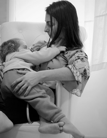 A tender moment between a woman and a young child as they cuddle in a comfortable chair. The woman holds the child close to her chest, and both appear to be relaxed and at peace. The scene is intimate and conveys a sense of warmth and connection.