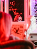 A mirror reflection showing a person's eyes and forehead, partially obscured by a brightly lit neon sign that reads 'Love You' with a heart symbol above it. The scene is bathed in red and pink hues, with a blurred foreground showing possibly a magazine or book.
