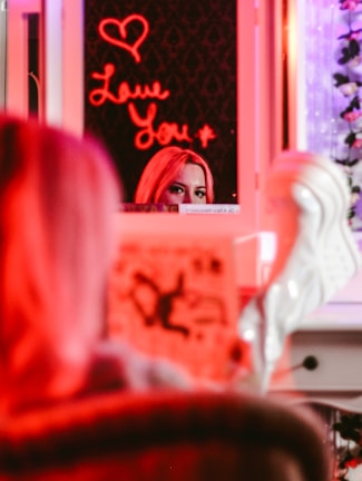 A mirror reflection showing a person's eyes and forehead, partially obscured by a brightly lit neon sign that reads 'Love You' with a heart symbol above it. The scene is bathed in red and pink hues, with a blurred foreground showing possibly a magazine or book.