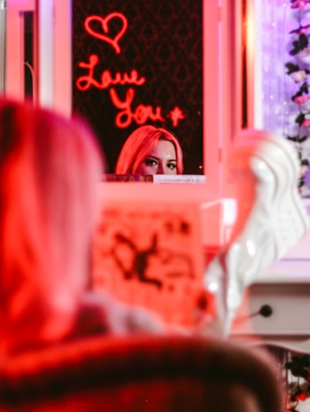 A mirror reflection showing a person's eyes and forehead, partially obscured by a brightly lit neon sign that reads 'Love You' with a heart symbol above it. The scene is bathed in red and pink hues, with a blurred foreground showing possibly a magazine or book.