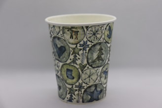 A paper cup decorated with a festive, winter-themed design featuring symbols such as a reindeer, snowflakes, a Christmas tree, a heart, and a star. The illustrations are colorful and arranged in a pattern that covers the entire cup. The color palette includes shades of green, blue, and gray, creating a cohesive and harmonious look.