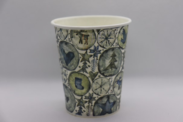 A paper cup decorated with a festive, winter-themed design featuring symbols such as a reindeer, snowflakes, a Christmas tree, a heart, and a star. The illustrations are colorful and arranged in a pattern that covers the entire cup. The color palette includes shades of green, blue, and gray, creating a cohesive and harmonious look.