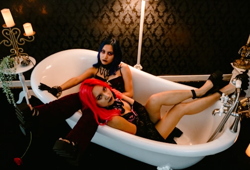 Two people with vibrant hair, one blue and one red, are sitting together in a white clawfoot bathtub. The scene is set in a dimly lit room with ornate wallpaper and candles providing a warm glow. The individuals have a striking appearance, dressed in dark, edgy clothing. A vintage white side table holds a small vase and a few candles.