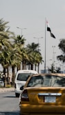 A yellow Hyundai sedan with a license plate is stationary on a palm-lined street. The background features another white vehicle and a national flag waving on a tall pole. Streetlights line the road under a partly cloudy sky.