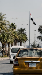 A yellow Hyundai sedan with a license plate is stationary on a palm-lined street. The background features another white vehicle and a national flag waving on a tall pole. Streetlights line the road under a partly cloudy sky.