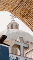 A stone church dome with a large cross on top is framed by a brick arch. The church features arched windows with shutters and a cross emblem on the sign below.