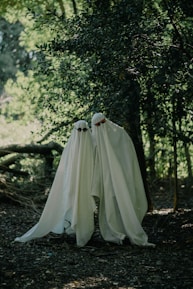 Two figures draped in white sheets resembling ghosts stand in a wooded area. They wear sunglasses, adding a whimsical touch to the eerie setting. The surrounding trees cast shadows, creating a mysterious and somewhat playful atmosphere.