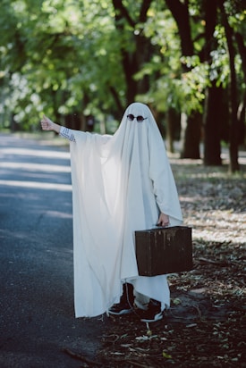 A person is dressed in a ghost costume made of a white sheet, complete with sunglasses, standing on the side of a deserted road. The individual is giving a thumbs-up gesture with one hand and holding a vintage suitcase in the other hand. The background features a tree-lined street with dappled sunlight filtering through the leaves.