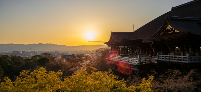 A traditional Japanese temple with ornate wooden architecture sits amidst lush trees and foliage under a clear sky, as the sun sets on the horizon casting a golden glow over the scene.