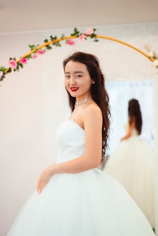 A woman wearing a white gown poses gracefully in front of a mirror, surrounded by a decorative floral arch with pink flowers. Her hair is styled in loose waves, and she is adorned with a choker necklace. The background is softly lit, creating an elegant and serene ambiance.