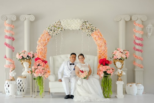 A couple dressed in elegant wedding attire is seated on a white tufted chair. The couple is surrounded by a beautiful arch adorned with an array of flowers, including white, peach, and pink blooms. The background features classical columns entwined with pink floral garlands, and ornate bust vases with floral arrangements are placed on either side.