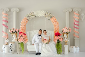 A couple dressed in elegant wedding attire is seated on a white tufted chair. The couple is surrounded by a beautiful arch adorned with an array of flowers, including white, peach, and pink blooms. The background features classical columns entwined with pink floral garlands, and ornate bust vases with floral arrangements are placed on either side.