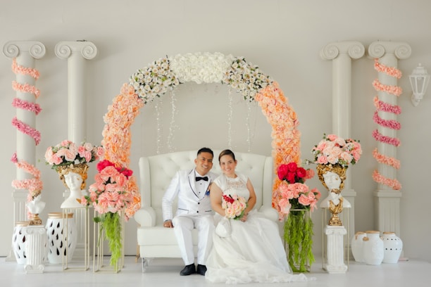 A couple dressed in elegant wedding attire is seated on a white tufted chair. The couple is surrounded by a beautiful arch adorned with an array of flowers, including white, peach, and pink blooms. The background features classical columns entwined with pink floral garlands, and ornate bust vases with floral arrangements are placed on either side.