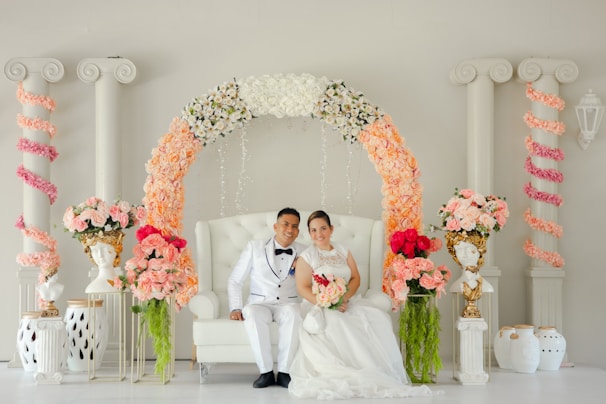 A couple dressed in elegant wedding attire is seated on a white tufted chair. The couple is surrounded by a beautiful arch adorned with an array of flowers, including white, peach, and pink blooms. The background features classical columns entwined with pink floral garlands, and ornate bust vases with floral arrangements are placed on either side.