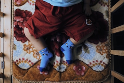 A child is lying on a patterned blanket with floral designs, wearing blue socks with cartoon faces and red shorts. The setting appears comfortable and cozy, suggesting a home environment.