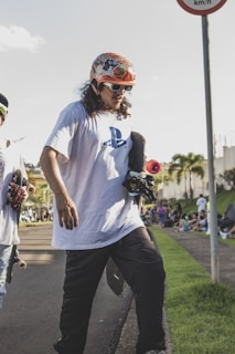 A person wearing a colorful helmet and sunglasses is holding a skateboard on a paved pathway. They are dressed in a white T-shirt with a PlayStation logo and dark pants. Another skateboarder is partially visible next to them. The scene is outdoors with a few palm trees in the background and a crowd gathered on the grass by the side.