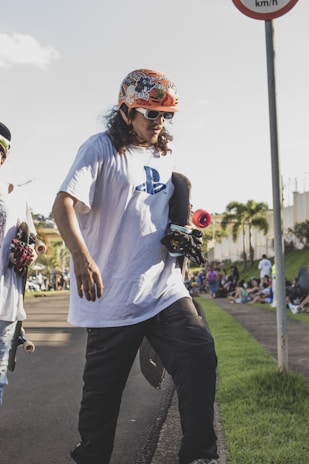 A person wearing a colorful helmet and sunglasses is holding a skateboard on a paved pathway. They are dressed in a white T-shirt with a PlayStation logo and dark pants. Another skateboarder is partially visible next to them. The scene is outdoors with a few palm trees in the background and a crowd gathered on the grass by the side.