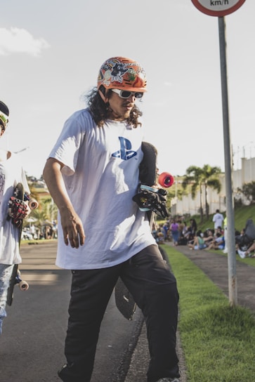 A person wearing a colorful helmet and sunglasses is holding a skateboard on a paved pathway. They are dressed in a white T-shirt with a PlayStation logo and dark pants. Another skateboarder is partially visible next to them. The scene is outdoors with a few palm trees in the background and a crowd gathered on the grass by the side.