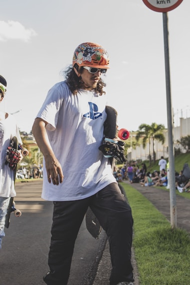 A person wearing a colorful helmet and sunglasses is holding a skateboard on a paved pathway. They are dressed in a white T-shirt with a PlayStation logo and dark pants. Another skateboarder is partially visible next to them. The scene is outdoors with a few palm trees in the background and a crowd gathered on the grass by the side.