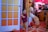 A child is standing next to a large, colorful door with a playful expression. The scene is decorated for a festive occasion with red and gold accents and a large musical instrument setup in the background. An adult in a white outfit is sitting nearby, looking at a phone.