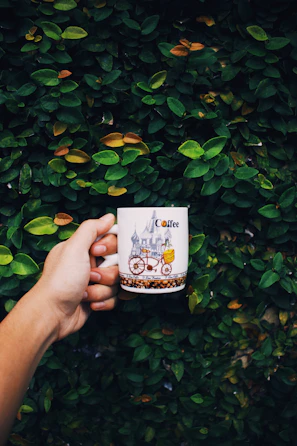 a hand holding a coffee mug in front of a hedge