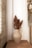 A textured ceramic vase containing dried flowers is placed on a wooden surface. The background features white fabric or curtains alongside a decorative arrangement of flowers and woven materials on the right. A textured brick wall is visible on the left, adding a rustic touch.