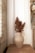 A textured ceramic vase containing dried flowers is placed on a wooden surface. The background features white fabric or curtains alongside a decorative arrangement of flowers and woven materials on the right. A textured brick wall is visible on the left, adding a rustic touch.
