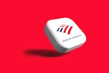 A white, rounded square object with the Bank of America logo displayed prominently. The logo features stylized red and blue stripes above the text 'BANK OF AMERICA'. The object casts a shadow on a vibrant red background, giving the impression that it is floating.