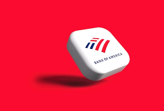 A white, rounded square object with the Bank of America logo displayed prominently. The logo features stylized red and blue stripes above the text 'BANK OF AMERICA'. The object casts a shadow on a vibrant red background, giving the impression that it is floating.