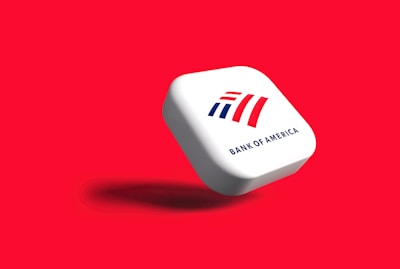 A white, rounded square object with the Bank of America logo displayed prominently. The logo features stylized red and blue stripes above the text 'BANK OF AMERICA'. The object casts a shadow on a vibrant red background, giving the impression that it is floating.