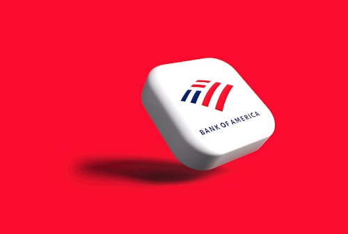 A white, rounded square object with the Bank of America logo displayed prominently. The logo features stylized red and blue stripes above the text 'BANK OF AMERICA'. The object casts a shadow on a vibrant red background, giving the impression that it is floating.