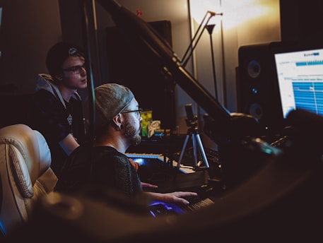 Two individuals are in a dimly lit recording studio, one seated and the other standing, intently focusing on a computer screen displaying audio editing software. The room contains professional audio equipment, including speakers and a microphone mounted on a stand. Ambient lighting creates a focused and creative atmosphere.