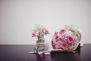 A bouquet of pink flowers and a small jar with pink roses and delicate white flowers are arranged on a dark wooden surface. The bouquet is wrapped in brown paper and includes a mix of roses, hydrangeas, and baby's breath. The jar is decorated with lace and twine, adding a rustic touch.