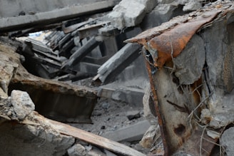 A scene of collapsed concrete structures with visible reinforcement steel bars and extensive rust. Debris and broken concrete slabs are scattered around, indicating a significant structural failure.