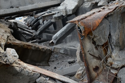 A scene of collapsed concrete structures with visible reinforcement steel bars and extensive rust. Debris and broken concrete slabs are scattered around, indicating a significant structural failure.