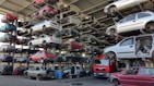 Numerous stacked rows of disassembled cars are placed on metal structures in what appears to be a scrapyard. Many of the cars are missing parts like wheels and doors.